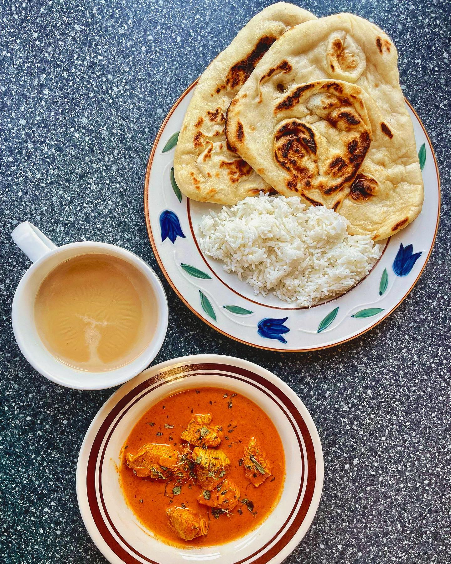 A cup of chai, plate of naan and rice, and a bowl of butter chicken on a table.